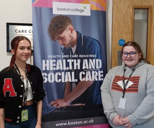 2 Health and Social Care Learners stood in front of Health and Social Care banner, looking directly into the camera whilst smiling