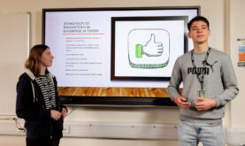 2 Boston College Business learners, presenting to the camera, using a presentation on the white board