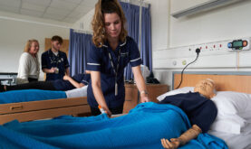 Health and Social Care Learner practicing on dummy in the college Care Core Ward. Another Leaner with lecturer in background