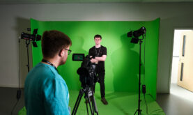 Boston College Learner stood infront of a Green Screen whilst another learner operatea a camera